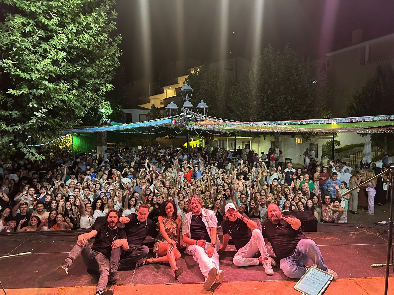 Banda de Por Camela sentada en el escenario con el público al fondo entre árboles