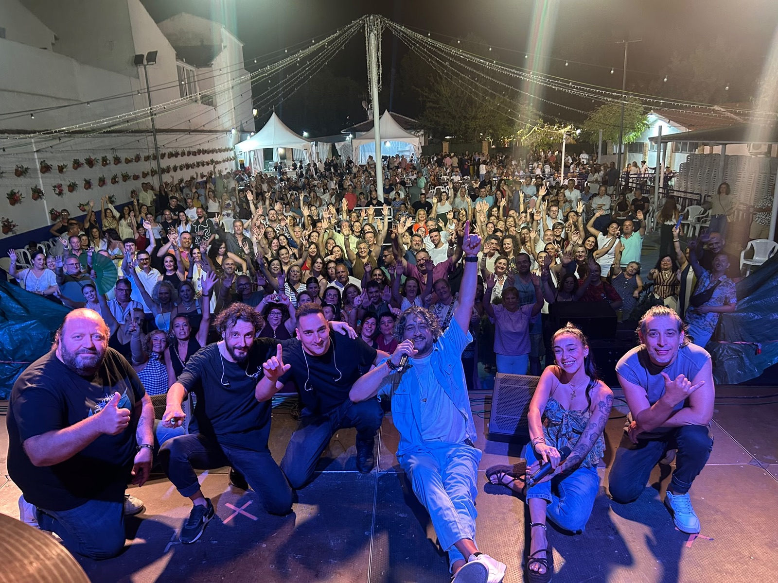 Banda de Por Camela arrodillada en el escenario con el público coreando en una feria de noche